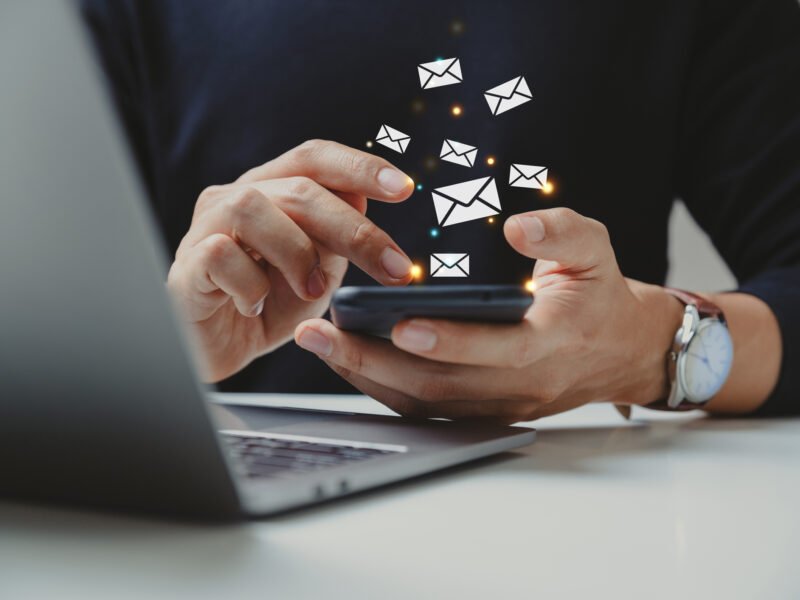 Hands using a smartphone with floating envelope icons beside a laptop, symbolizing email marketing.