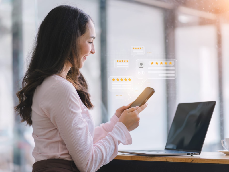 Woman at a desk using her smartphone while positive five-star customer review notifications appear, with a laptop and coffee beside her in a bright office setting.
